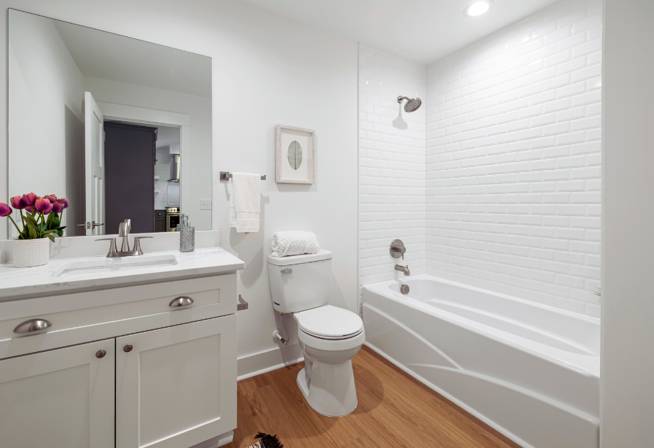 view of small white bathroom with tub on the far right, toilet is centered in the photo to the left of the bathtub.  to the left of the toilet is a small single sink with a mirror on the wall and flowers on the countertop.   
